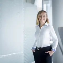 Uśmiechnięta kobieta w białej koszuli i czarnych spodniach z ozdobnym paskiem, stojąca w nowoczesnym wnętrzu.