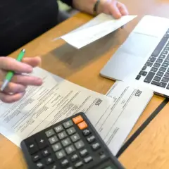 Dłoń z długopisem nad dokumentami ZUS, obok kalkulator i laptop.