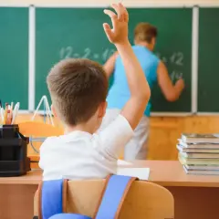Uczeń z podniesioną ręką przed tablicą, na której nauczyciel pisze.