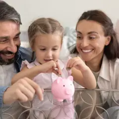 Rodzina uczy córkę oszczędzania. Dziewczynka wkłada monetę do różowej świnki-skarbonki, rodzice patrzą z uśmiechem. To początek budowania kapitału na przyszłość, może nawet na 1000 plus.