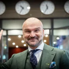 Rafał Brzoska, z uśmiechem i uniesionym palcem, symbolizuje swój rosnący majątek. Za nim zegary pokazują różne strefy czasowe.