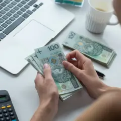 Dłonie trzymające banknoty 100 zł, obok laptop, kalkulator i okulary.