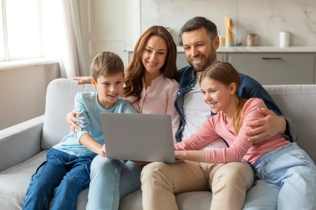 Rodzina ogląda coś na laptopie, śmiejąc się. Dzieci siedzą na kolanach rodziców. To wspaniałe chwile, warte więcej niż 1000 plusów.