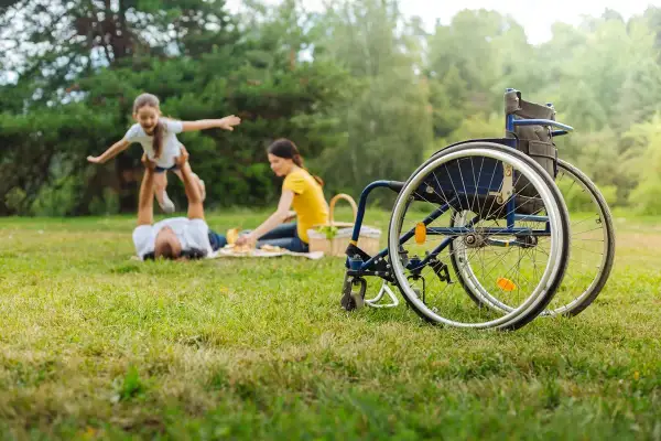 Rodzina na pikniku. Ojciec podrzuca córkę, matka przygotowuje jedzenie. Obok stoi wózek inwalidzki, symbol wsparcia i obecności opiekuna osoby niepełnosprawnej.