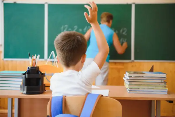 Uczeń z podniesioną ręką przed tablicą, na której nauczyciel pisze.