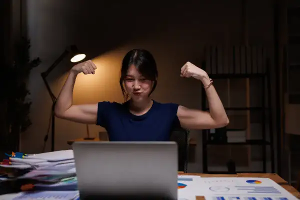 Młoda kobieta w nocy przy biurku pokazuje bicepsy, jakby cieszyła się z dodatku za pracę w nocy.