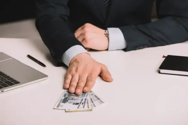 Dłoń mężczyzny w garniturze podaje banknoty rubli na białym biurku obok laptopa.