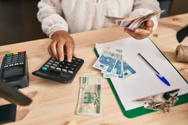 Kobieta liczy pieniądze na kalkulatorze, przygotowując się do rozliczeń związanych z samozatrudnieniem. Na stole leżą polskie banknoty.
