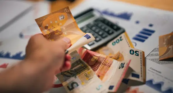 Dłoń trzyma banknot 50 euro nad stosem innych banknotów i kalkulatorem. To symbol prowizji i finansów.
