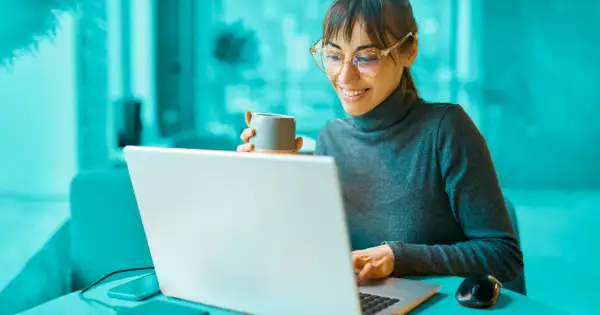 Uśmiechnięta kobieta z kubkiem kawy przegląda na laptopie informacje o dofinansowania na otwarcie firmy.