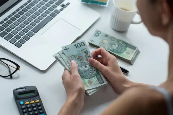 Dłonie trzymające banknoty 100 zł, obok laptop, kalkulator i okulary.